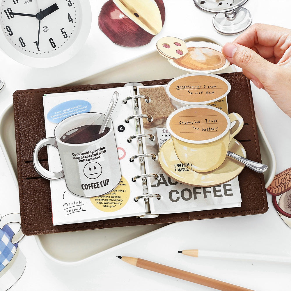 Hand holding coffeethemed paper stickers featuring cups saucers and notes displayed on a brown leather planner