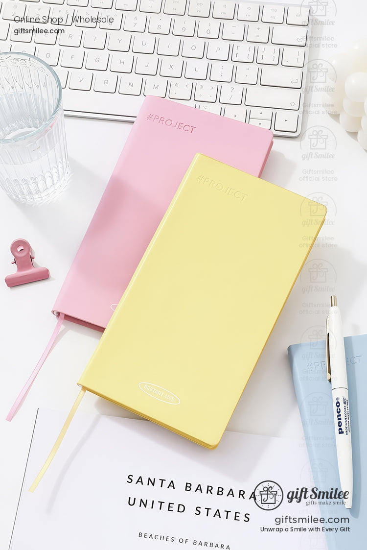 Pastel yellow and pink hardcover project notebooks on a white desk with keyboard, pen, and glass — minimalist stationery flat lay for productivity and gifting.