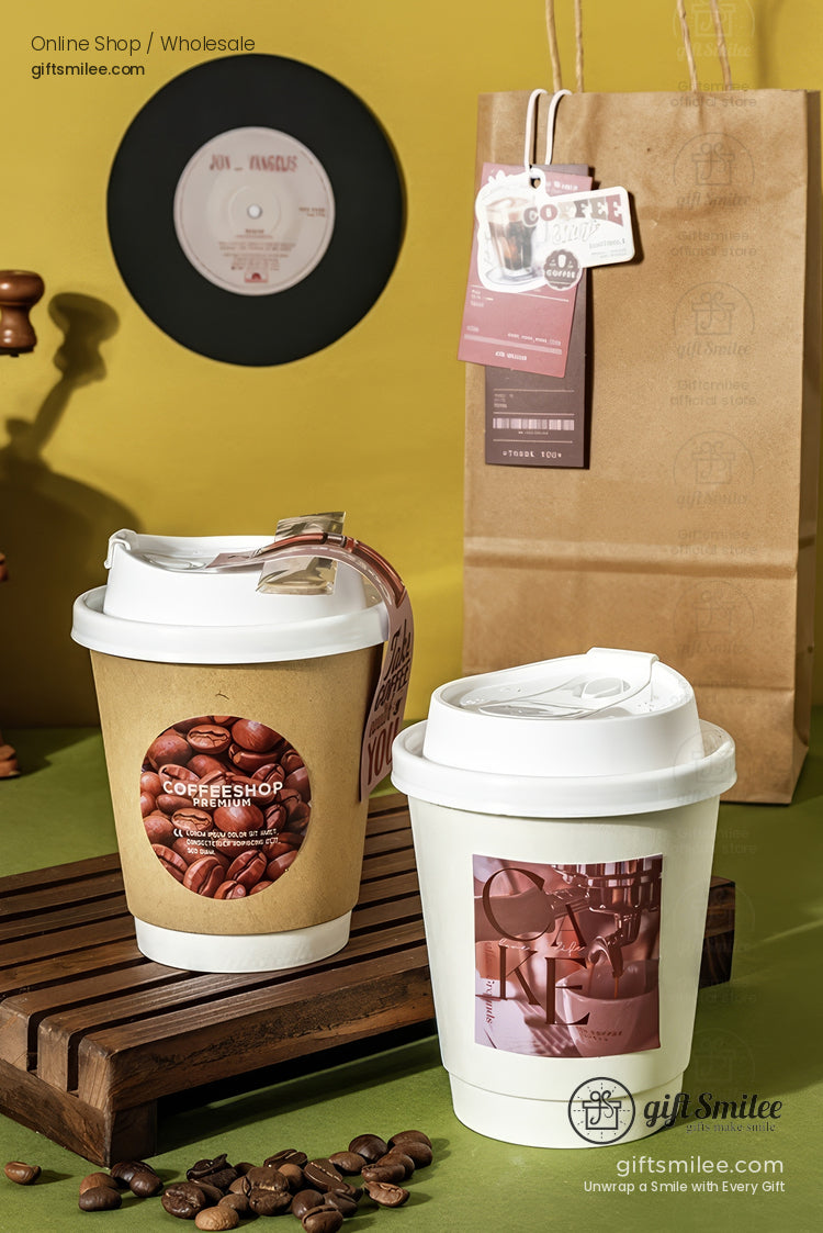 Brown paper coffee cup with coffee bean print and white lid next to a white cup on a wooden tray with beans scattered