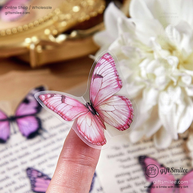 Pink translucent butterfly sticker with detailed wings displayed on a fingertip against floral and vintage book decor